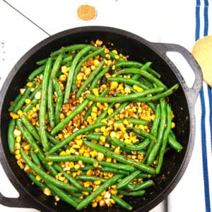 Cast iron skillet filled with corn and green beans on a white surface with a wood spoon, blue and white napkin, and measuring spoons of spices around it.