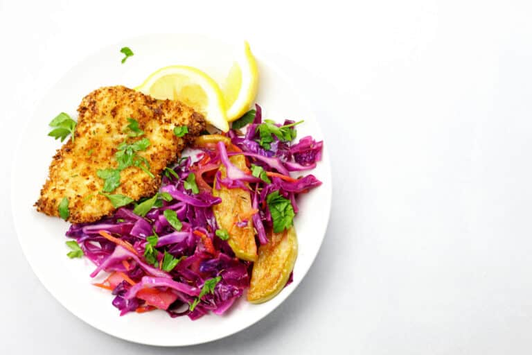 A white plate with a serving of breaded and fried chicken breast, garnished with chopped herbs and accompanied by roasted potato wedges and a colorful salad of red cabbage and mixed vegetables. Lemon wedges are also on the plate.