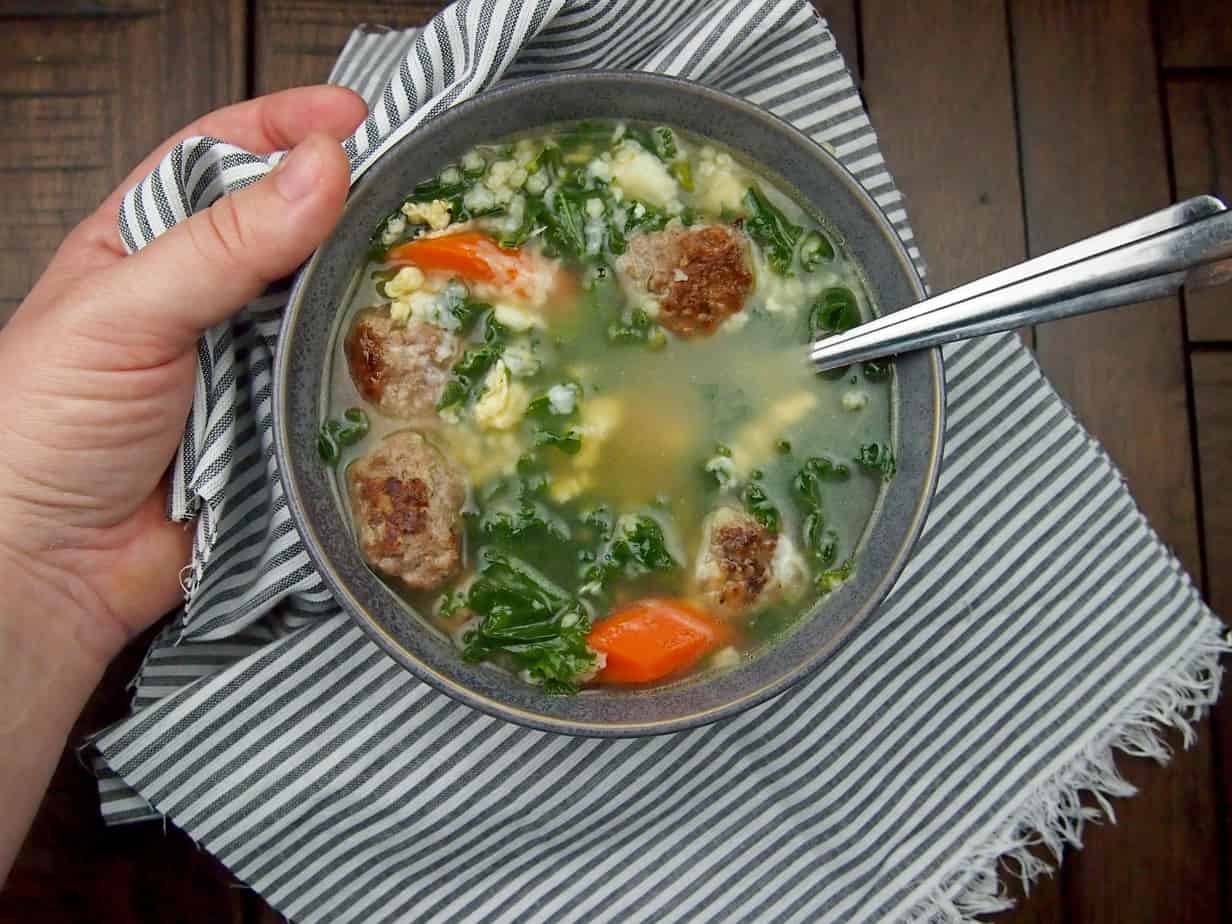 Holding a bowl of Italian wedding soup with meatballs, carrots, onions, and kale