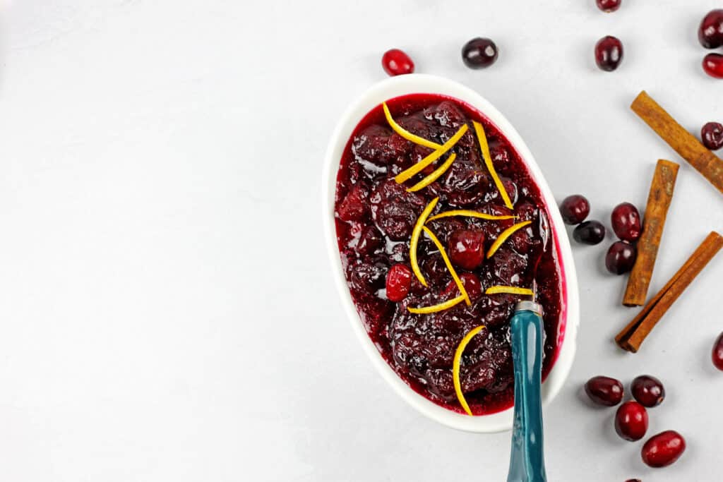 The best ever cranberry sauce in a white bowl on a white background with fresh cranberries scattered next to it.