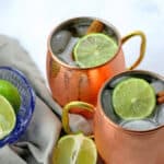 Two copper mugs of Bourbon mules on a white countertop garnished with limes and a blue crystal bowl of limes on the side. A gray napkin is under the bowl.