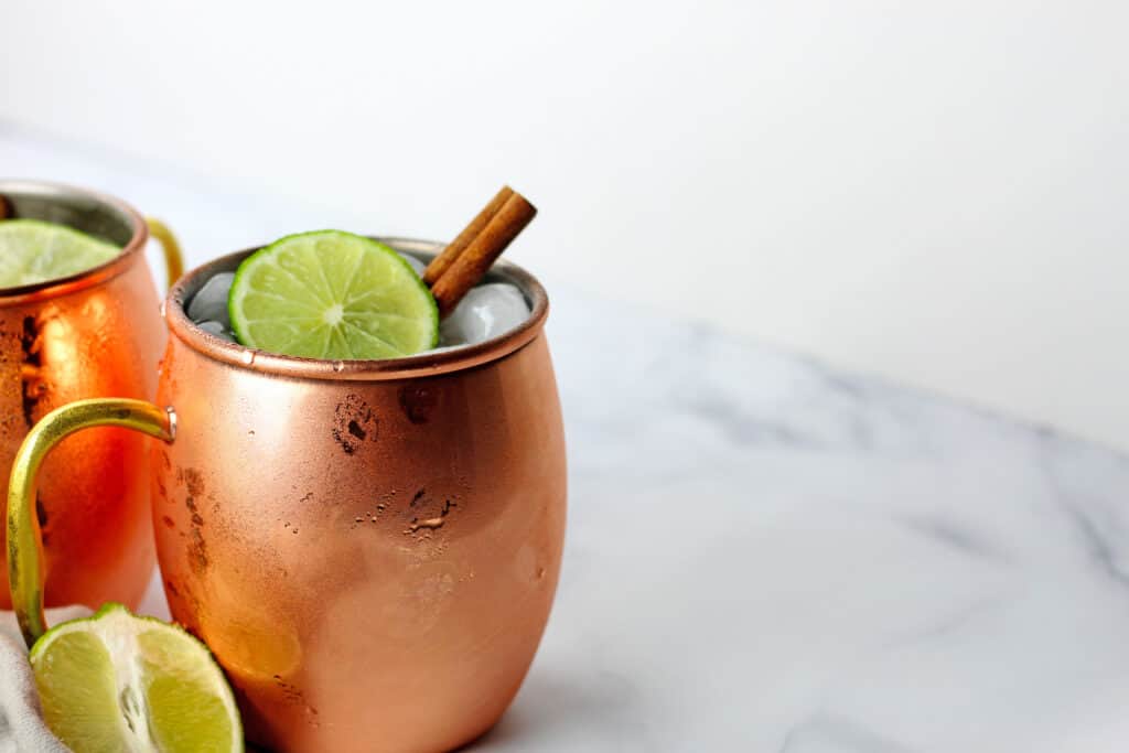 One copper mug of a bourbon mule garnished with a lime and cinnamon stick. Extra lime is next to the mug.