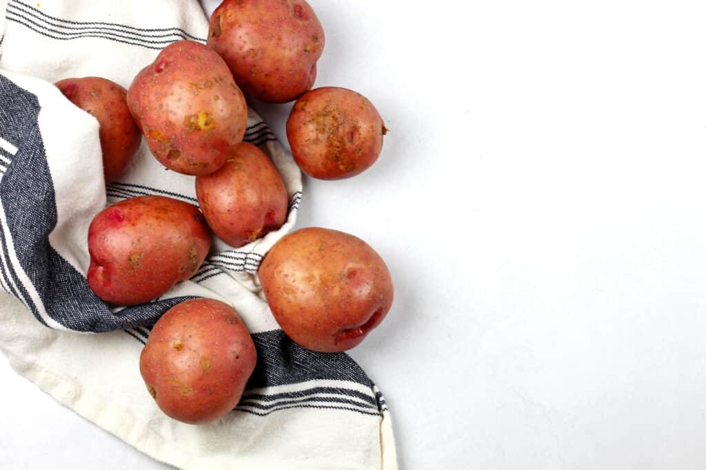 Whole red skin potatoes ready to be made into red skin mashed potatoes on a white surface with a blue and white tea towel