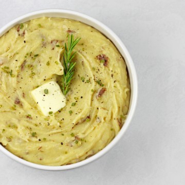 Overview of red skin mashed potatoes with a pat of melting butter and sprig of rosemary on top on a white surface.