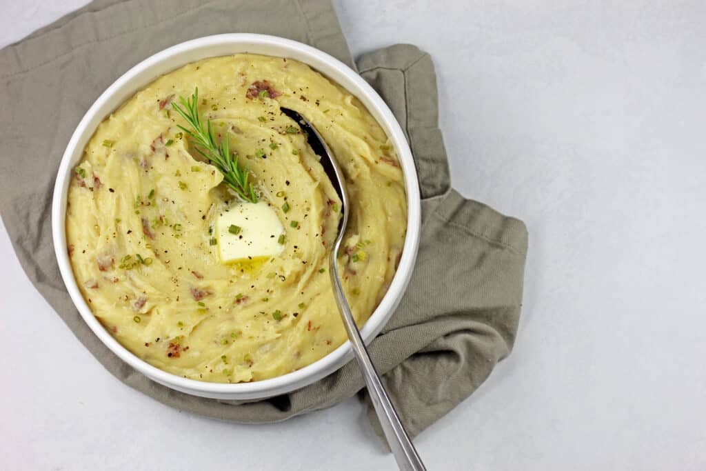 Overview of red skin mashed potatoes with a pat of melting butter, sprig of rosemary, and a serving spoon on top on a white surface with a gray towel.