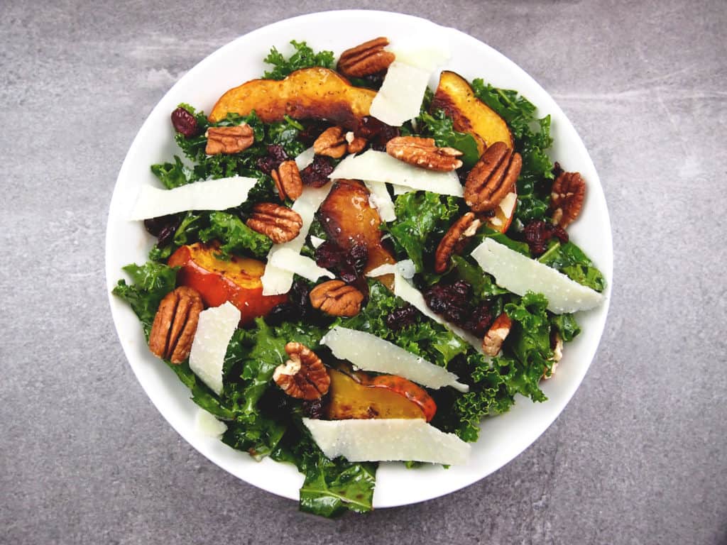 Overview of roasted acorn squash on top of massaged kale salad with pecans, shaved Parmesan, and dried cranberries in a bowl