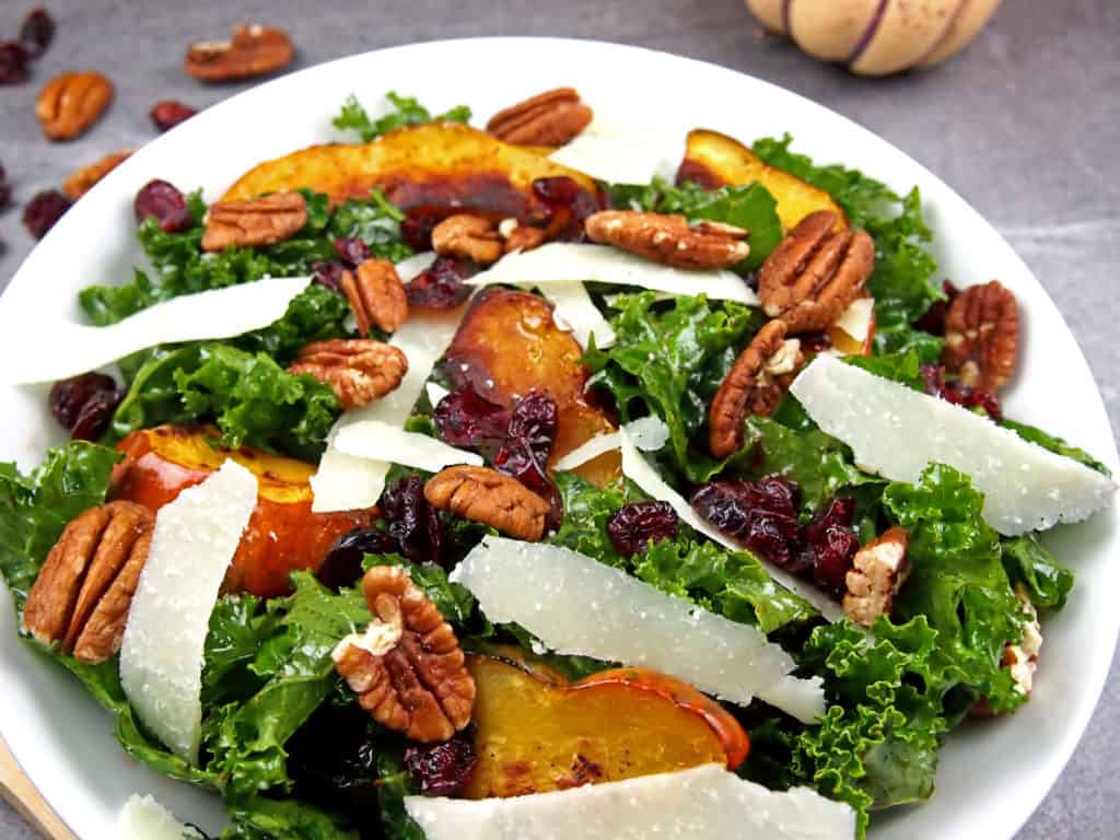 Close up of roasted acorn squash on top of massaged kale salad with pecans, shaved Parmesan, and dried cranberries in a bowl