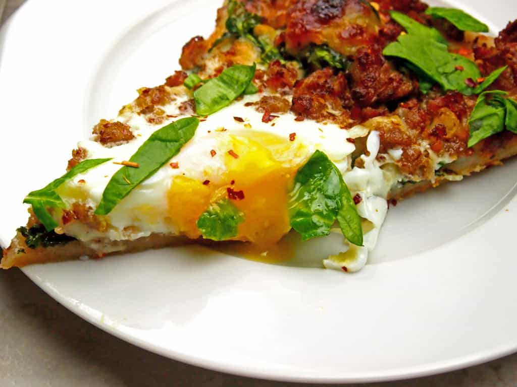 Close up of pizza slice with drippy egg, breakfast sausage, and spinach on top.