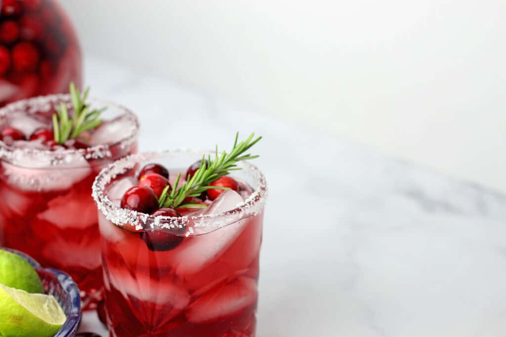 Two mistletoe margaritas situated on the left with limes on the side and a pitcher of mistletoe margaritas in the background and atop of a white marble countertop.