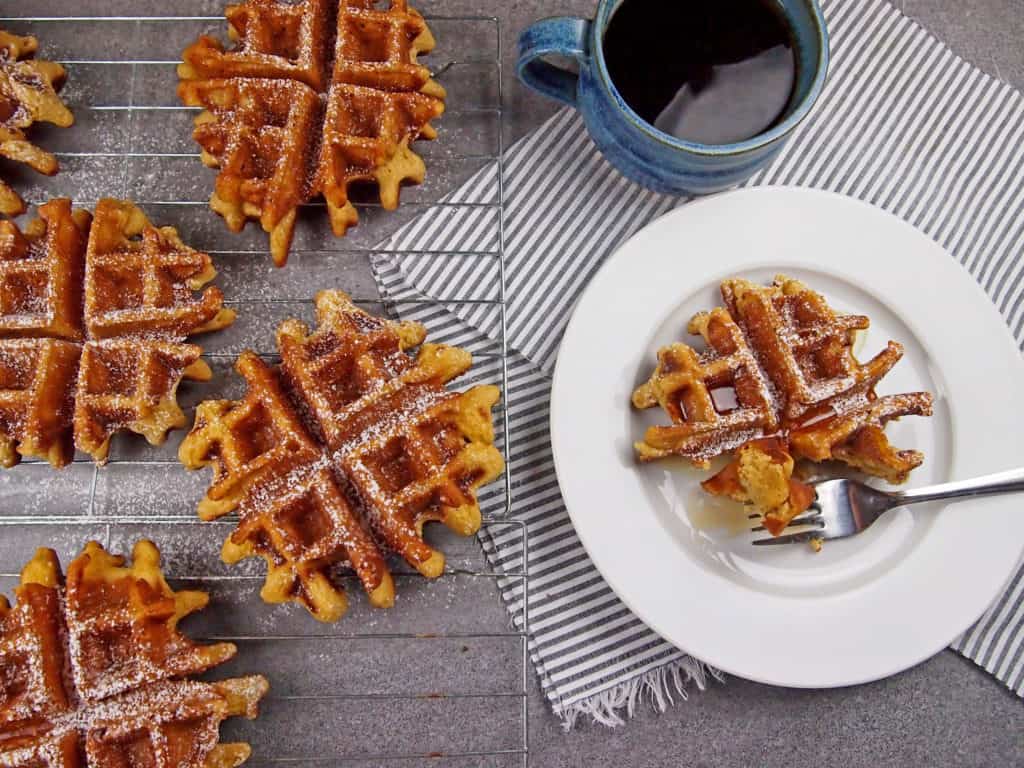 View of eggnog waffles on a cooling rack next to a plate of waffles with syrup and coffee on the side