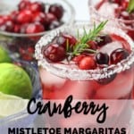A picture of a bowl of limes and cranberries next to two mistletoe margaritas with salted rims and fresh cranberries and rosemary as garnishes on top of a white countertop. The text overlay says "cranberry mistletoe margaritas".