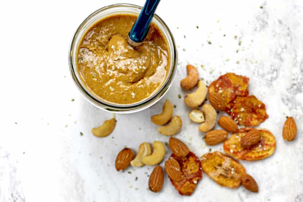 Overview of banana nut butter with a spoon in the jar with nuts and dried bananas scattered on a white background. 