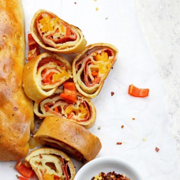 Sliced pepperoni rolls with one intact pepperoni roll sitting on a white background with red pepper flakes and bell peppers scattered.