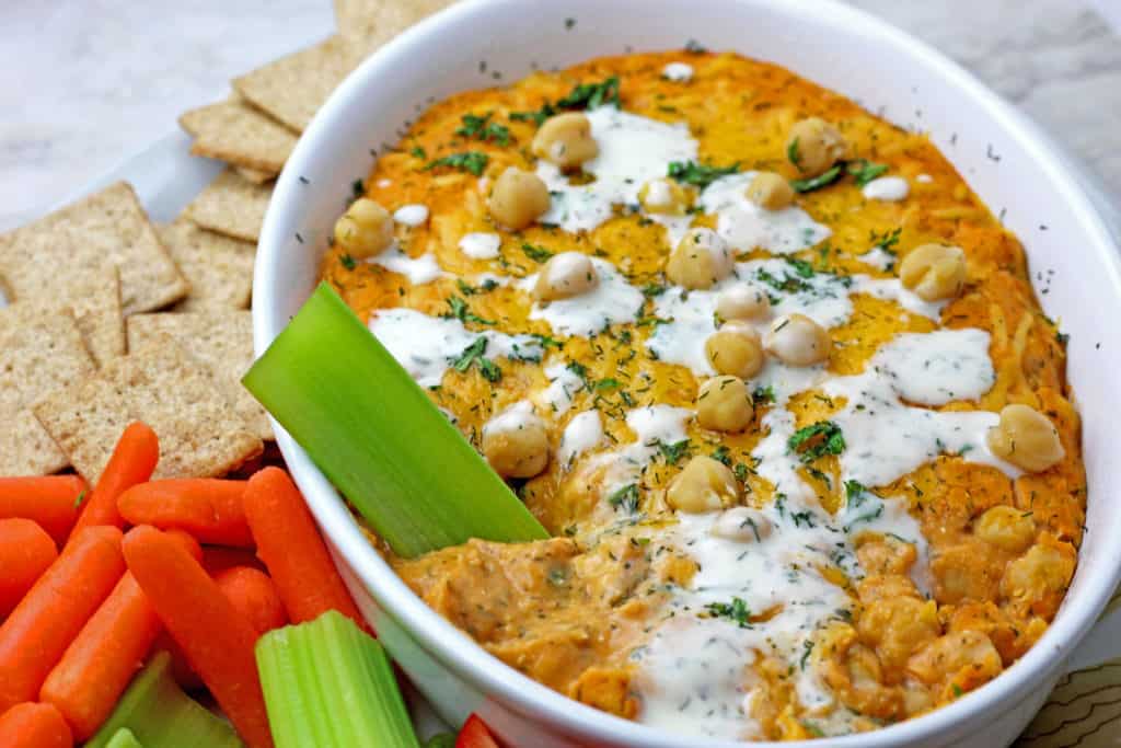 Buffalo chickpea dip with piece of celery dipped in surrounded by veggies and crackers on a plate.