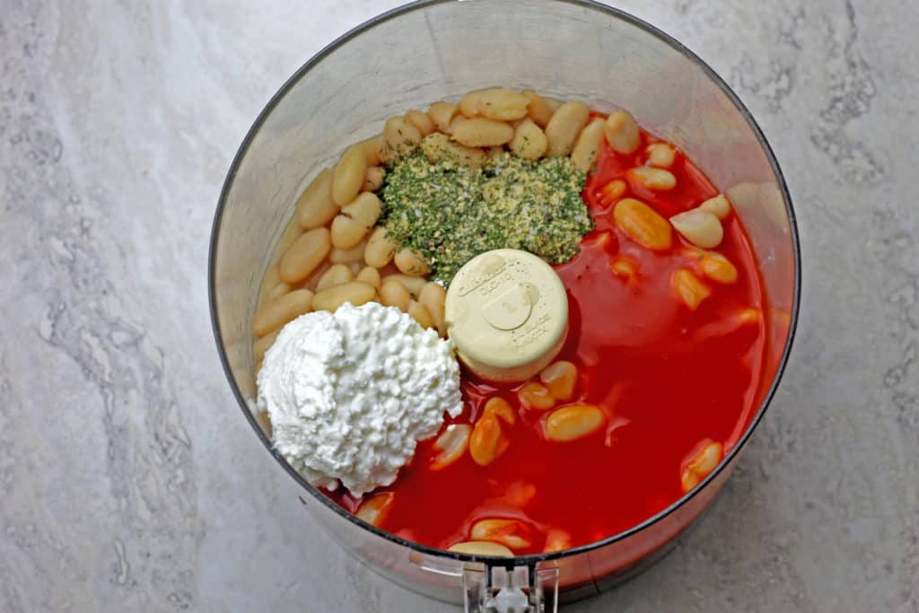 Ingredients for buffalo chickpea dip in a food processor bowl (cottage cheese, buffalo sauce, seasonings, and beans).