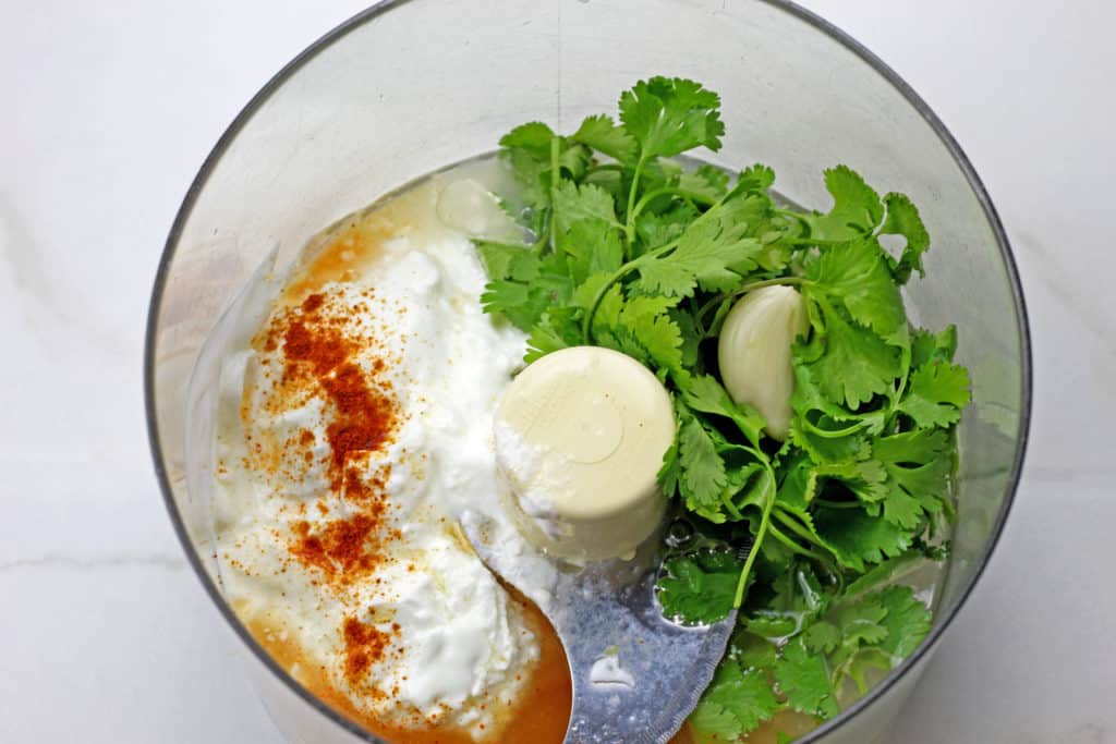 Food processor bowl with cilantro, lime juice, cayenne pepper, yogurt, and garlic inside the bowl getting ready to be made into cilantro garlic sauce.