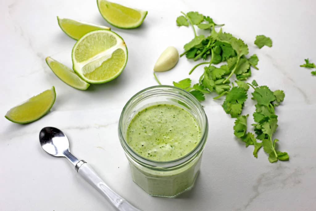 Jar of cilantro garlic sauce surrounding by a spoon, limes, garlic clove, and cilantro on a white counter.