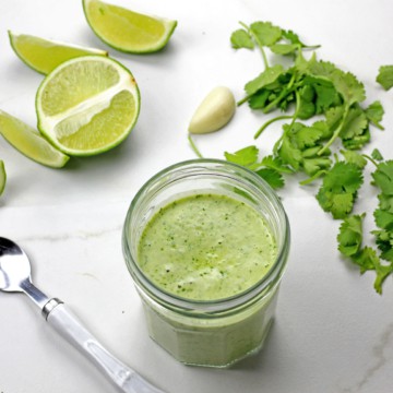 Jar of cilantro garlic sauce surrounding by a spoon, limes, garlic clove, and cilantro on a white counter.