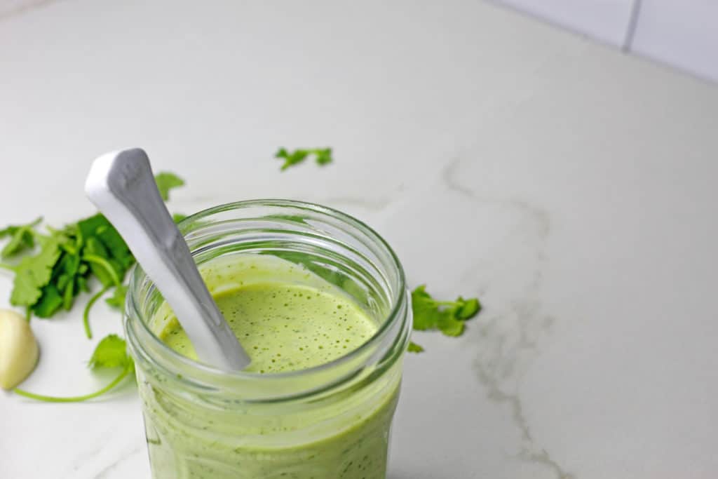 Jar of cilantro garlic sauce with a spoon in the jar and cilantro in the background on a white counter.