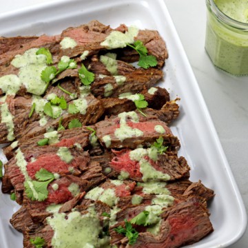 Platter of instant pot carne asada with cilantro garlic sauce drizzled on top and a jar of the sauce on the side.