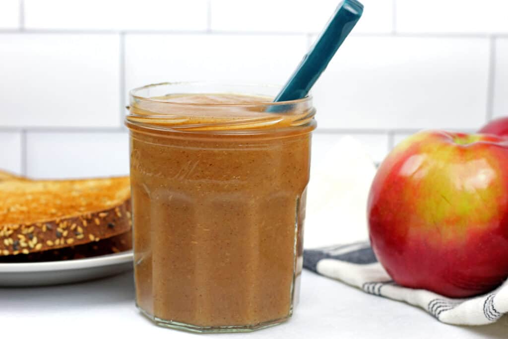 Jar of instant pot apple butter with a spoon resting in the jar with toast and whole apples on the side on a white background.