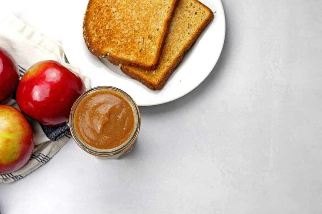 Overview from above of a jar of instant pot apple butter with whole apples and toast on the side on a white-gray background.