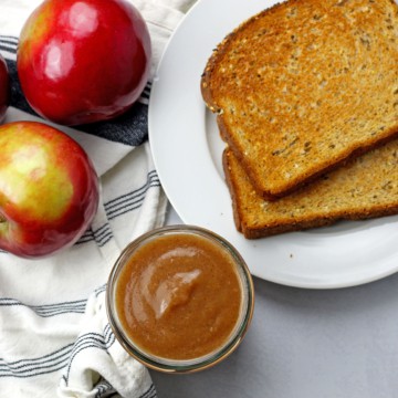 Overview from above of a jar of instant pot apple butter with whole apples and toast on the side on a white-gray background.