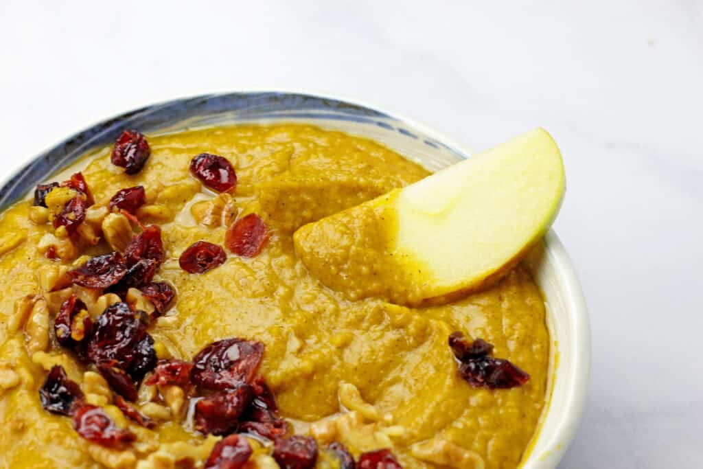 Close up of pumpkin pie hummus with dried cranberries, crushed nuts, and an apple dipped in the hummus.