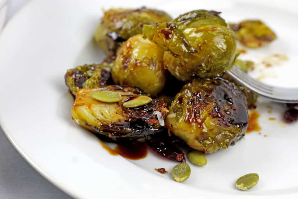 Close up of smashed brussels sprouts with balsamic glaze, pepitas, and cranberries on a white plate.