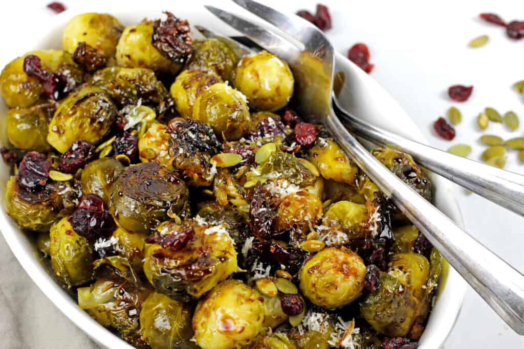 Close up of smashed brussels sprouts in a bowl with pepitas, cranberries and balsamic glaze with serving spoons.