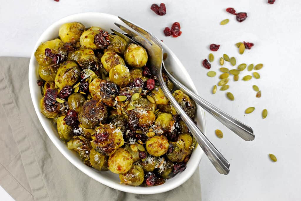 Smashed brussels sprouts in a bowl with pepitas, cranberries and balsamic glaze with serving spoons.