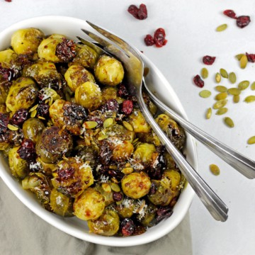 Smashed brussels sprouts in a bowl with pepitas, cranberries and balsamic glaze with serving spoons.