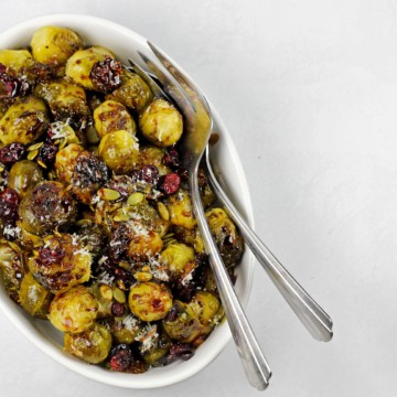 Smashed brussels sprouts in a bowl with pepitas, cranberries and balsamic glaze with serving spoons on a white background.