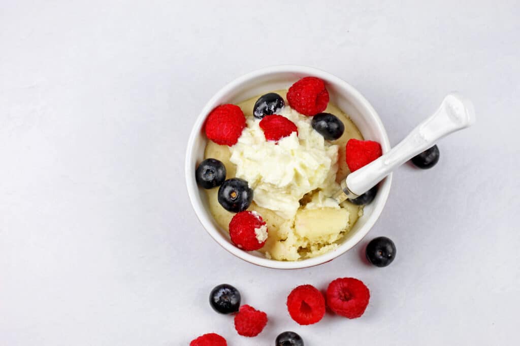 Overview of the vanilla mug cake with a white spoon on a white surface with whipped cream and mixed berries.