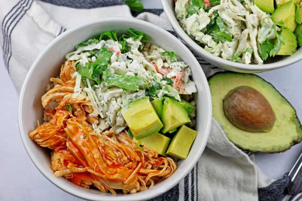 Close up of two buffalo chicken rice bowl with an avocado and a napkin on the side on a white background.