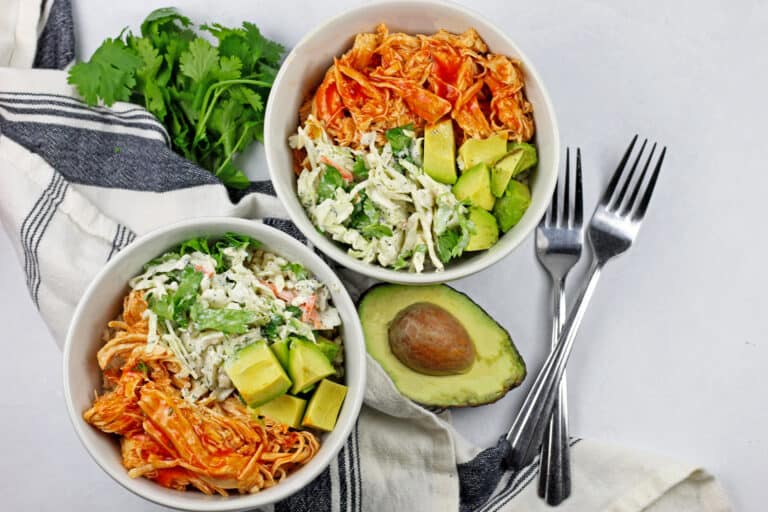 overview of two buffalo chicken rice bowl with an avocado, cilantro, forks, and a napkin on the side on a white background.