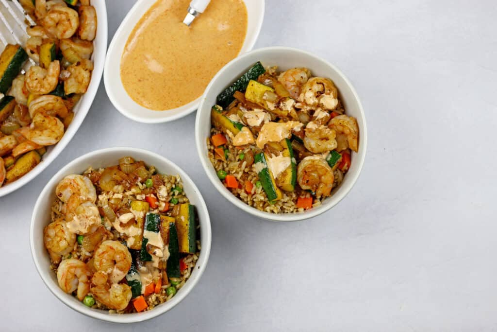 Two hibachi shrimp rice bowls on a white background and in white bowls. On the side there is a pan of shrimp and vegetables as well as yum yum sauce with a white spoon.
