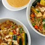 Two hibachi shrimp rice bowls on a white background and in white bowls. On the side there is a pan of shrimp and vegetables as well as yum yum sauce with a white spoon. With the text overlay saying "hibachi shrimp bowls"