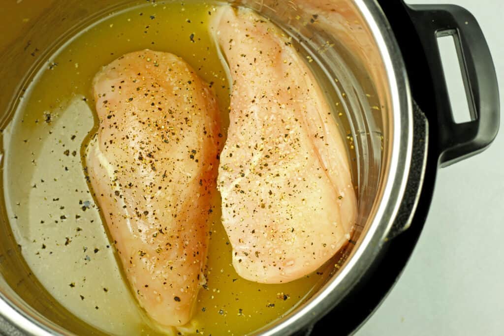 View of two raw chicken breasts in chicken broth with seasonings in an instant pot to make instant pot shredded chicken