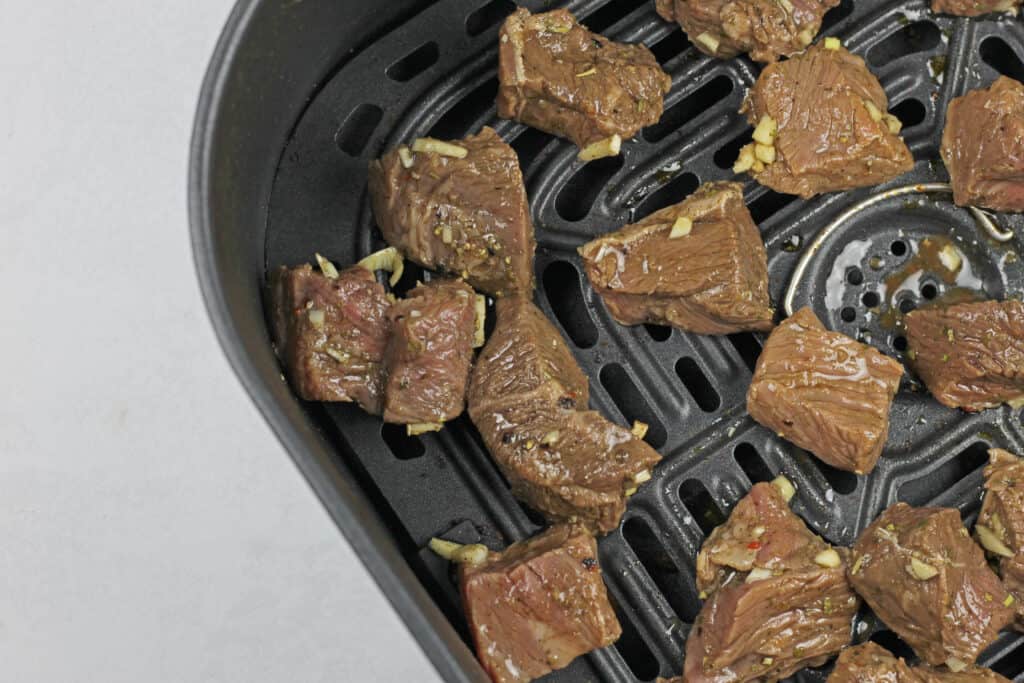 Raw steak bites in a single layer in a black air fryer basket.