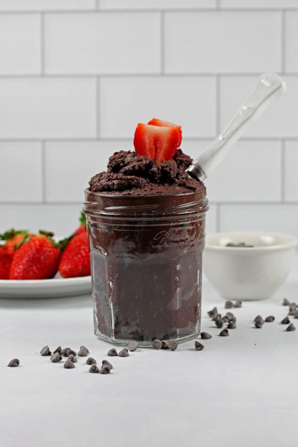A jar full of edible brownie batter with a white spoon and chocolate chips and strawberries on the side on a white subway tile background.