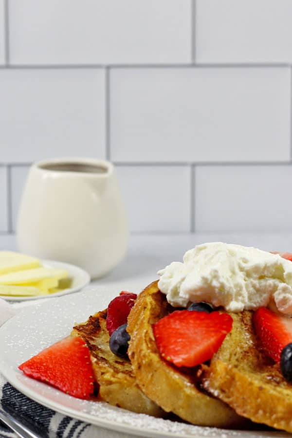 A white plate of sourdough french toast with berries and yogurt on top with a side of butter and maple syrup.