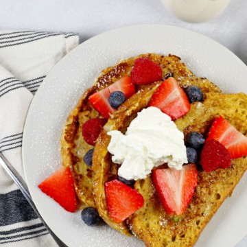 A plate of sourdough french toast with berries and yogurt on top with a side of butter and maple syrup on a white background. There is a fork and blue and white napkin on the side also.