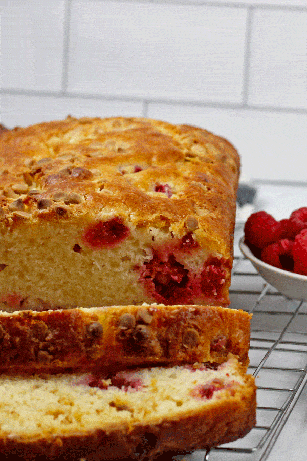 The White Chocolate and Raspberry Loaf Cake on a cooling rack sliced with a small bowl of raspberries on the side with subway tile in the background.