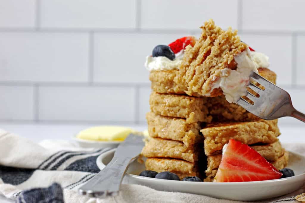 A fork with a bite of gluten free oat flour pancakes taken out of a stack of pancakes. The pancakes are topped with yogurt and berries. On side side there is a butter knife, a butter dish and a blue and white napkin. The background is a white subway tile backsplash.