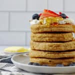 A stack of gluten free oat flour pancakes with yogurt and berries on top. Maple syrup is dripping down the sides of the pancakes. On side there is a napkin and a butter dish with white subway tile in the background.