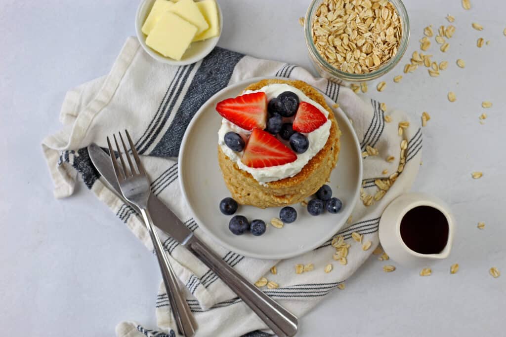 A plate of gluten free oat flour pancakes topped with berries and yogurt with butter, oats, syrup, a blue and white napkin, and silverware on the side on a white background.