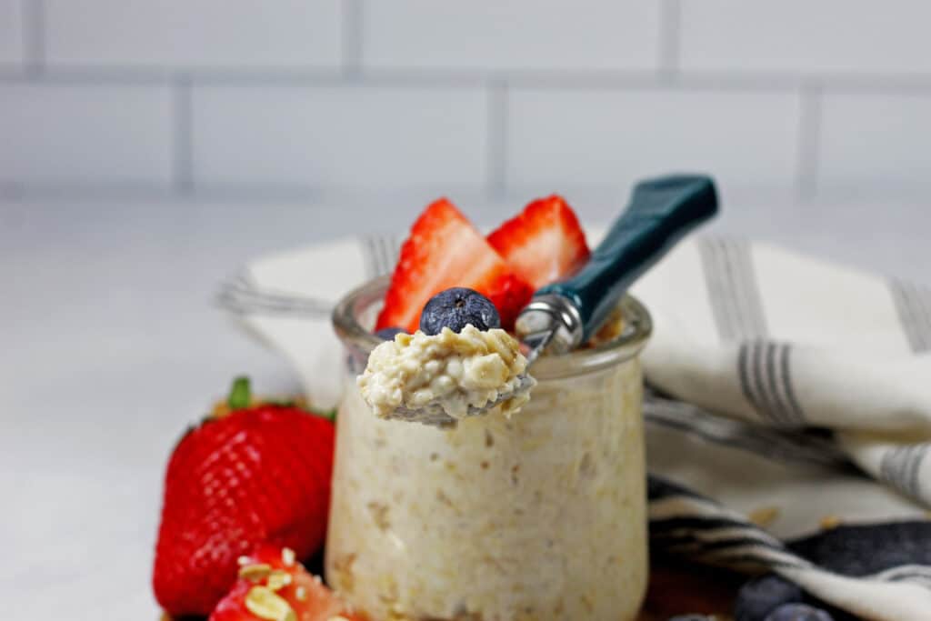 A jar of vanilla overnight oats with berries on a top and on the side on a wooden cutting board and a white and blue napkin on the side. White subway tile is in the background. A spoon is resting on top of the jar with oats and a blueberry on the spoon.
