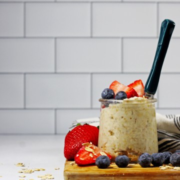 A jar of vanilla overnight oats with fruit and a spoon on a wooden cutter board and a white and blue napkin along with white subway tile in the background.