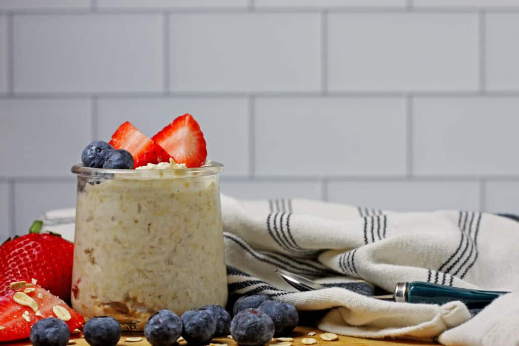 A jar of vanilla overnight oats with berries on a top and on the side on a wooden cutting board and a white and blue napkin on the side. White subway tile is in the background.